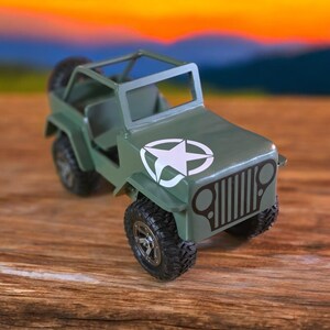 K&ouml;nnte beinhalten: Ein gr&uuml;ner Spielzeug-Jeep mit einem wei&szlig;en Stern-Emblem auf der Motorhaube. Der Jeep hat schwarze Reifen, einen &Uuml;berrollk&auml;fig und ein Reserverad hinten. Der Hintergrund ist ein verschwommenes Bild eines Sonnenuntergangs.