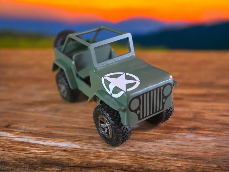 K&ouml;nnte beinhalten: Ein gr&uuml;ner Spielzeug-Jeep mit einem wei&szlig;en Stern-Emblem auf der Motorhaube. Der Jeep hat schwarze Reifen, einen &Uuml;berrollk&auml;fig und ein Reserverad hinten. Der Hintergrund ist ein verschwommenes Bild eines Sonnenuntergangs.
