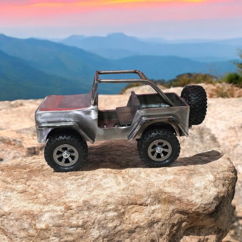 K&ouml;nnte beinhalten: Ein kleines Spielzeug-Jeep aus Metall mit einem Ersatzreifen auf der R&uuml;ckseite. Der Jeep hat ein rotes Dach und steht auf einer felsigen Oberfl&auml;che mit einem Bergpanorama. Die Reifen sind schwarz und die R&auml;der sind silberfarben.