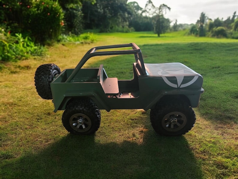 K&ouml;nnte beinhalten: Ein gr&uuml;ner Spielzeug-Jeep mit schwarzen Reifen und einem &Uuml;berrollb&uuml;gel. Der Jeep hat ein Ersatzrad hinten und eine wei&szlig;e Grafik auf der Motorhaube. Der Jeep steht auf einer Wiese.