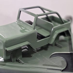 K&ouml;nnte beinhalten: Ein kleines, olivgr&uuml;nes Spielzeug-Jeep mit &Uuml;berrollk&auml;fig, Sitzen und detaillierter Karosserie. Das Modell hat eine matte Oberfl&auml;che und &auml;hnelt einem klassischen Gel&auml;ndewagen. Das Spielzeug ist zum Spielen oder Ausstellen geeignet.