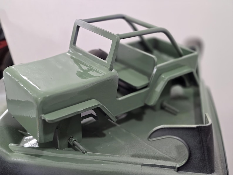 K&ouml;nnte beinhalten: Ein kleines, olivgr&uuml;nes Spielzeug-Jeep mit &Uuml;berrollk&auml;fig, Sitzen und detaillierter Karosserie. Das Modell hat eine matte Oberfl&auml;che und &auml;hnelt einem klassischen Gel&auml;ndewagen. Das Spielzeug ist zum Spielen oder Ausstellen geeignet.