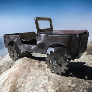 K&ouml;nnte beinhalten: Metallskulptur eines Jeeps mit dunkler, strukturierter Oberfl&auml;che. Das Fahrzeug hat zahnradf&ouml;rmige R&auml;der und ein offenes Design. Es steht auf einer felsigen Oberfl&auml;che, mit Bergen und blauem Himmel im Hintergrund.