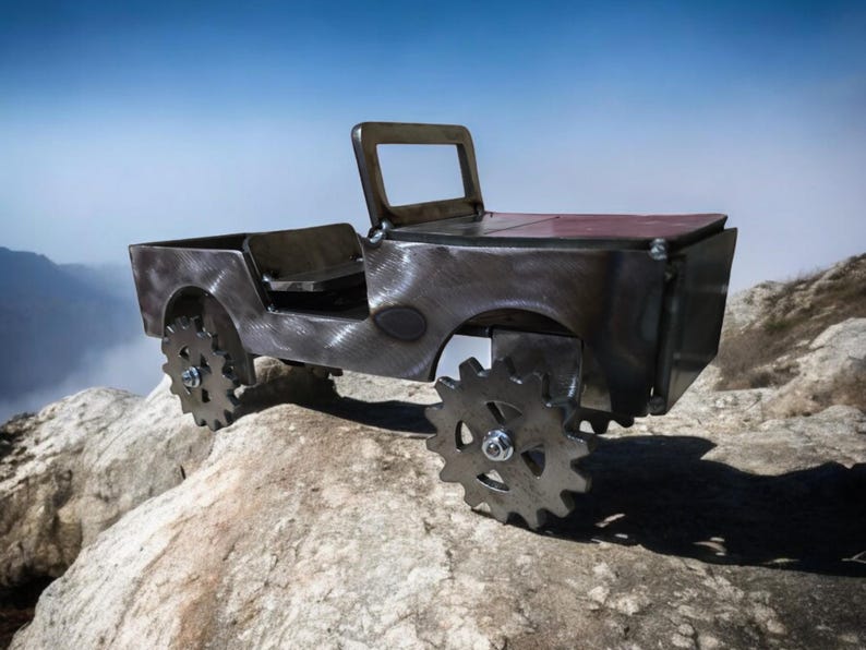 K&ouml;nnte beinhalten: Metallskulptur eines Jeeps mit dunkler, strukturierter Oberfl&auml;che. Das Fahrzeug hat zahnradf&ouml;rmige R&auml;der und ein offenes Design. Es steht auf einer felsigen Oberfl&auml;che, mit Bergen und blauem Himmel im Hintergrund.