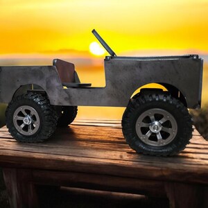 K&ouml;nnte beinhalten: Ein handgefertigtes Metall-Spielzeug-Jeep mit gro&szlig;en, robusten Reifen und einem braunen Sitz. Der Jeep hat eine Windschutzscheibe und einen &Uuml;berrollb&uuml;gel. Das Spielzeug steht auf einem Holztisch mit Sonnenuntergang im Hintergrund.