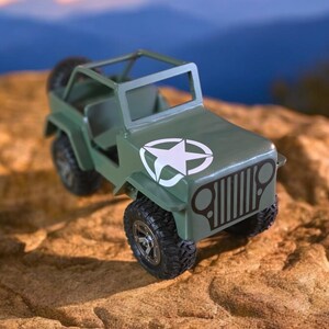 K&ouml;nnte beinhalten: Ein gr&uuml;ner Spielzeug-Jeep mit einem wei&szlig;en Stern-Emblem auf der Motorhaube. Das Miniaturfahrzeug hat schwarze Reifen und einen &Uuml;berrollk&auml;fig. Der Jeep steht vor einer verschwommenen Landschaft und einer strukturierten, beigefarbenen Oberfl&auml;che.
