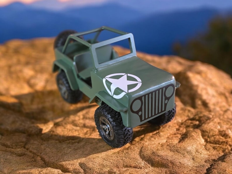 K&ouml;nnte beinhalten: Ein gr&uuml;ner Spielzeug-Jeep mit einem wei&szlig;en Stern-Emblem auf der Motorhaube. Das Miniaturfahrzeug hat schwarze Reifen und einen &Uuml;berrollk&auml;fig. Der Jeep steht vor einer verschwommenen Landschaft und einer strukturierten, beigefarbenen Oberfl&auml;che.