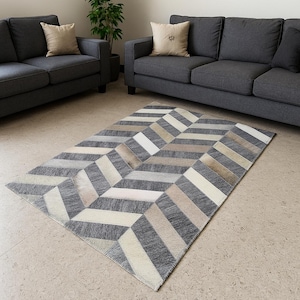 Könnte beinhalten: Rechteckiger Teppich mit geometrischem Zickzackmuster. Er hat abwechselnde Streifen in Grau, Weiß und Beige. Der Teppich liegt auf einem hell gefliesten Boden, mit zwei grauen Sofas und einer Pflanze im Hintergrund.