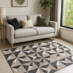 May include: A geometric patterned rug in grey, beige, and white tones. A light grey sofa with cushions is against a neutral wall. An abstract painting in a gold frame and a small side table with a lamp complete the room's decor.