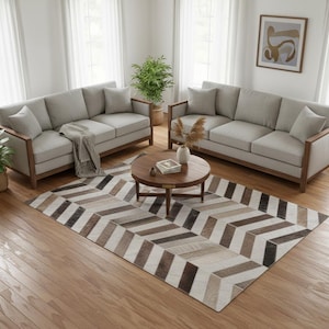 Peut inclure: Un salon avec deux canapés gris clair avec des cadres en bois, une table basse ronde et un tapis à motifs. Le tapis présente un motif en chevrons dans des tons de marron, beige et noir. La pièce est lumineuse avec la lumière naturelle.