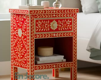 May include: Red bedside table with floral and leaf design. The table has a drawer and an open shelf. The table is next to a bed with white sheets and a light green duvet. A window is in the background.