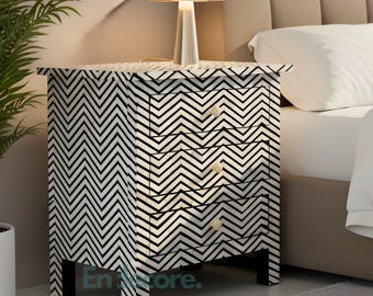 May include: A black and white chevron patterned nightstand with three drawers, a small lamp, and a potted plant. The nightstand is next to a bed with white bedding. The room has a neutral color palette.