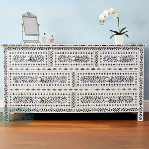 May include: A white chest of drawers with six drawers, decorated with a navy blue floral and geometric pattern. It has a mirror, perfume bottles, and an orchid in a pot on top. The room has a light blue wall and a wooden floor.