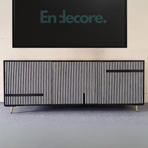 Op de afbeelding: Een zwart televisiescherm toont de tekst "En decore." boven een zwart-wit gestreepte kast. De kast heeft goudkleurige poten en een minimalistisch ontwerp. De achtergrond is een lichtbeige muur en een grijze vloer.