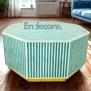 May include: An octagonal coffee table with a green and white striped design. The table has a yellow base and the text "En decore." on the top. The table is in a living room with a blue sofa and hardwood flooring.