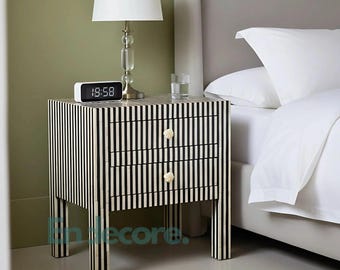 May include: A black and white striped nightstand with two drawers and gold-colored knobs. A white lamp with a clear glass base and a digital clock displaying 19:58 sit on top. The nightstand is next to a bed with white linens.
