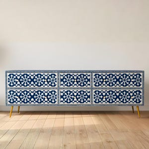 May include: A low, long chest of drawers with a navy blue and white geometric pattern. The six drawers sit on gold-coloured legs. A potted plant is to the left of the chest. The chest is approximately 150 cm long.