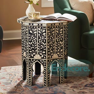 May include: A black and white hexagonal side table with a floral design. The table has a decorative pattern and arched legs. A cup and saucer, a vase with flowers, and an open book rest on the table. The table is approximately 24 inches tall.