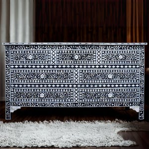 May include: A black and white dresser with floral and geometric inlay patterns. The dresser has six drawers with silver knobs. The piece is set on a dark wood floor with a white rug in front.