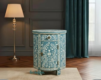 May include: A teal-colored, round bedside cabinet with a floral inlay design. The cabinet has a single drawer and a door, both adorned with brass hardware. A matching floor lamp with a cream-colored lampshade stands nearby.