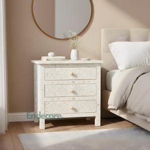 Può includere: Un comodino bianco con tre cassetti, ornato da un motivo floreale. Il tavolo ha pomelli colorati chiari ed è posto accanto a un letto con un cuscino bianco e biancheria da letto beige. Uno specchio rotondo è appeso alla parete sopra il tavolo.