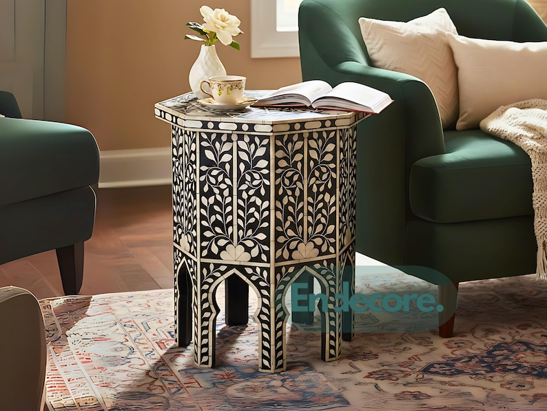 May include: A black and white hexagonal side table with a floral design. The table has a decorative pattern and arched legs. A cup and saucer, a vase with flowers, and an open book rest on the table. The table is approximately 24 inches tall.