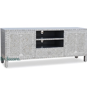 May include: Gray wooden TV stand with floral inlay design. The cabinet has two doors, a drawer, and open shelving. The stand is rectangular and has four short legs. The doors and drawer feature silver hardware.