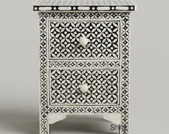 May include: A rectangular bedside table featuring two drawers with a black and white geometric floral pattern. The piece has a flat top surface with a border design and two round white knobs centered on each drawer front, supported by four feet.