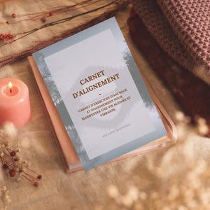Puede incluir: Un diario rosa titulado "CARNET D'ALIGNEMENT" con texto en francés, incluyendo "CARNET D'EXERCICES D'ANCRAGE ET D'ALIGNEMENT POUR MANIFESTER UNE VIE ALIGNÉE ET VIBRANTE." Una vela rosa encendida y flores secas en el fondo.