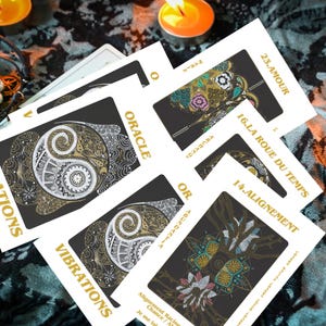 Peut inclure: Un jeu de cartes oracle avec des motifs complexes en or, blanc et noir. Les cartes présentent divers symboles et textes, dont "Oracle", "Vibrations" et des numéros. Les cartes sont disposées sur une surface à motifs, avec des bougies allumées en arrière-plan.