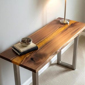 May include: A wooden console table with a live edge design and a yellow resin inlay. The table has a rectangular top and metal legs. A lamp with a white shade sits on the table, along with a bowl and books.