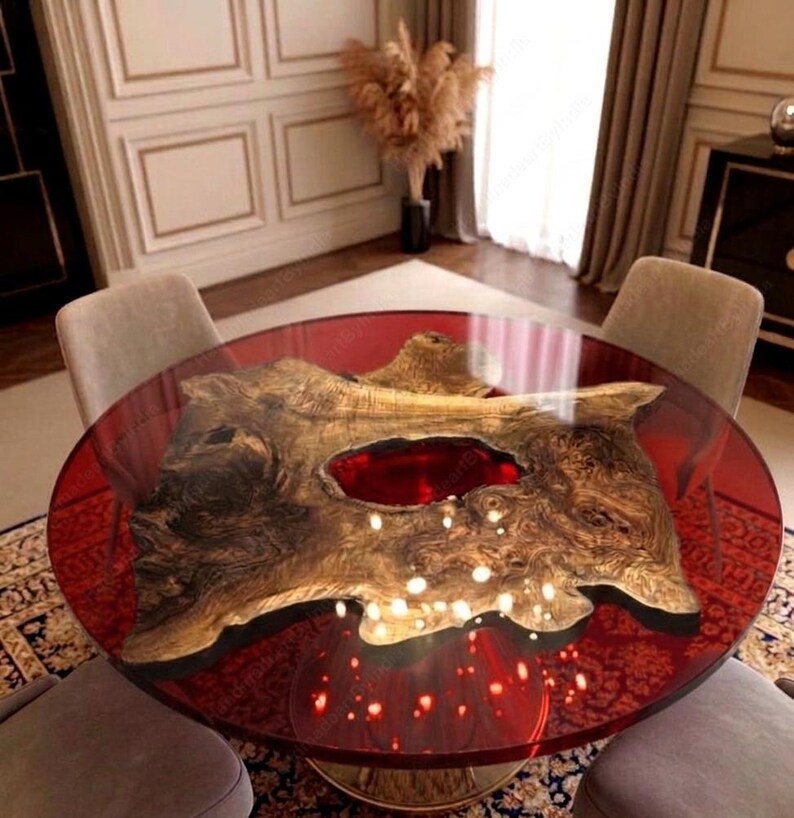 May include: A round dining table with a red translucent top and a natural wood centerpiece. The wood has a unique shape and a reddish-brown hue. The table is supported by a gold-colored base and is surrounded by four gray chairs.