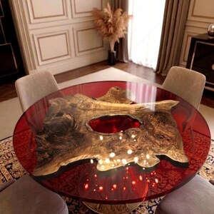 May include: A round dining table with a red translucent top and a natural wood centerpiece. The wood has a unique shape and a reddish-brown hue. The table is supported by a gold-colored base and is surrounded by four gray chairs.