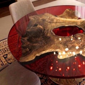 May include: A round table with a clear, red-tinted resin top. The table features a natural wood centerpiece with a large, red-filled void. The table is supported by a gold-colored base. The table's diameter is approximately 48 inches.