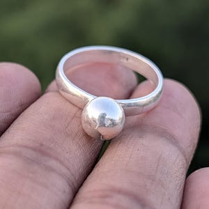 Puede incluir: Un anillo de plata con una banda lisa y redondeada y una prominente cuenta esférica. El anillo se muestra en un primer plano, destacando su superficie pulida y su diseño simple. El anillo se sostiene en una mano sobre un fondo verde borroso.