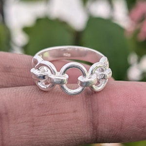Peut inclure: Une bague en argent avec un motif de maillons de chaîne. La bague comporte trois maillons de chaîne reliés, avec un maillon circulaire au centre. La bague est présentée sur un fond flou de feuillage vert et rose. C'est un bijou.