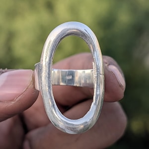 May include: A silver ring with an oval-shaped design. The ring is held up against a blurred green background. The ring's band is a simple, rectangular shape.