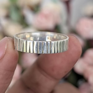 Puede incluir: Un anillo plateado con un diseño de líneas verticales texturizadas. El anillo se sostiene en una mano, con un fondo borroso de flores rosas y blancas. La superficie del anillo tiene una serie de líneas paralelas grabadas.