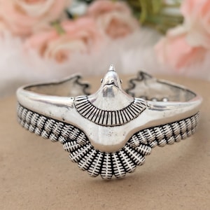 May include: Silver cuff bracelet with an eagle motif. The bracelet showcases detailed feather and wing patterns, with an eagle head at the centre. The background features blurred pink flowers.