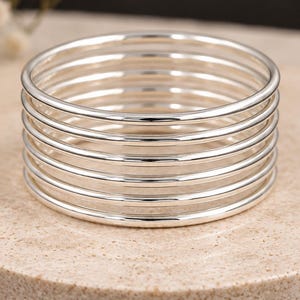 Puede incluir: Una pila de siete brazaletes finos y pulidos de color plateado. Cada brazalete es una banda circular lisa con un acabado metálico reflectante. Los brazaletes están dispuestos verticalmente en una sola columna, creando un aspecto uniforme.