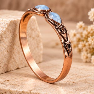 Puede incluir: Una pulsera de metal en tono cobre con dos piedras cabujón de color azul claro en forma ovalada, engastadas en biseles de metal decorativos y texturizados. La banda muestra intrincados patrones de volutas en relieve.