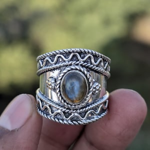 May include: A silver ring with an oval, iridescent gemstone at the center. The ring features intricate rope and floral designs around the band. The gemstone appears to have blue and green hues.