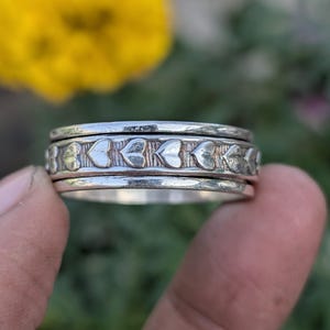 Puede incluir: Un anillo de plata con un diseño de corazón. El anillo tiene una banda texturizada con formas de corazón repetidas. El anillo se sostiene entre los dedos sobre un fondo borroso amarillo y verde.