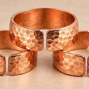 Pode incluir: Três anéis abertos feitos de cobre polido, cada um apresentando uma textura martelada distinta na superfície externa. Os anéis estão dispostos em uma formação triangular, exibindo o brilho metálico e o design arredondado consistente das bandas.