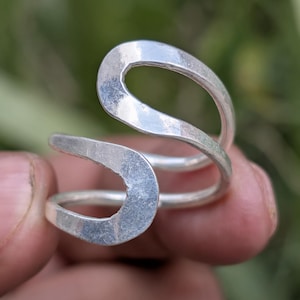 Op de afbeelding: Een zilverkleurige ring met een uniek, open ontwerp. De ring heeft een gehamerde textuur en een vloeiende, gebogen vorm. Het ontwerp van de ring wikkelt zich om de vinger en creëert een moderne en artistieke uitstraling. De ring wordt tegen een vage groene achtergrond gehouden.