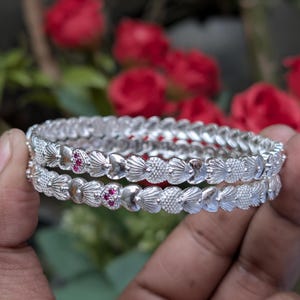 Può includere: Due braccialetti d'argento con intricati disegni floreali e a forma di cuore, impreziositi da piccole gemme rosse. I braccialetti sono tenuti in una mano, con uno sfondo sfocato di fiori rossi e fogliame verde.