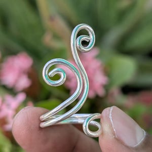 Può includere: Un anello color argento con un design unico a spirale. L'anello presenta due spirali prominenti e una fascia che avvolge il dito. L'anello è mostrato su uno sfondo di fogliame verde e fiori rosa.