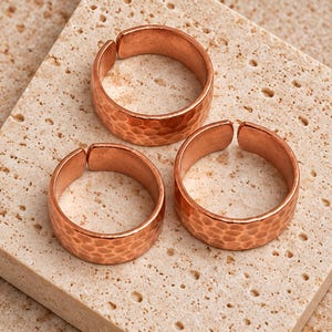 May include: Three copper rings with an open band design and a hammered texture. Each ring features a metallic reddish-brown finish and a wide, circular shape with a small gap at the back, showcasing a consistent reflective surface throughout.
