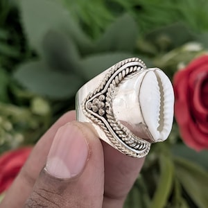 Puede incluir: Un anillo de plata con una gran concha de cauri blanca como pieza central. El anillo tiene detalles intrincados alrededor de la concha, con un diseño trenzado y detalles florales. El anillo se muestra sobre un fondo de follaje verde y una rosa roja.