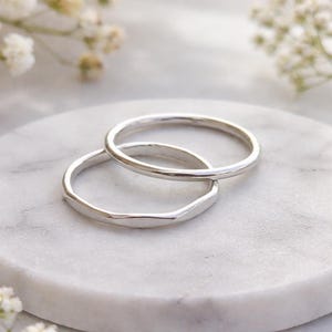 May include: Two silver rings, one with a smooth band and the other with a textured, irregular band. The rings are displayed on a white marble surface, with soft focus white flowers in the background. The rings are likely made of sterling silver.
